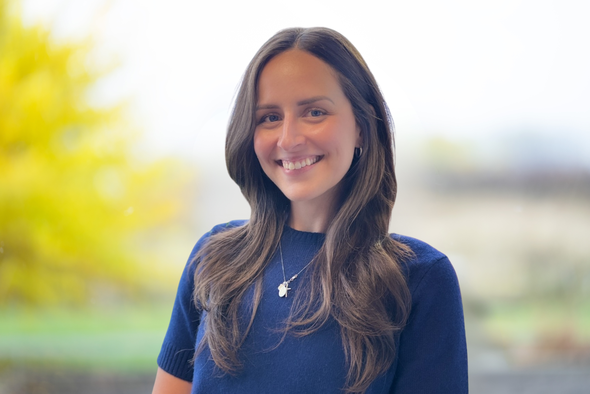 Martina with long dark hair, wearing a white top, standing outdoors with blurred yellow trees in the background.