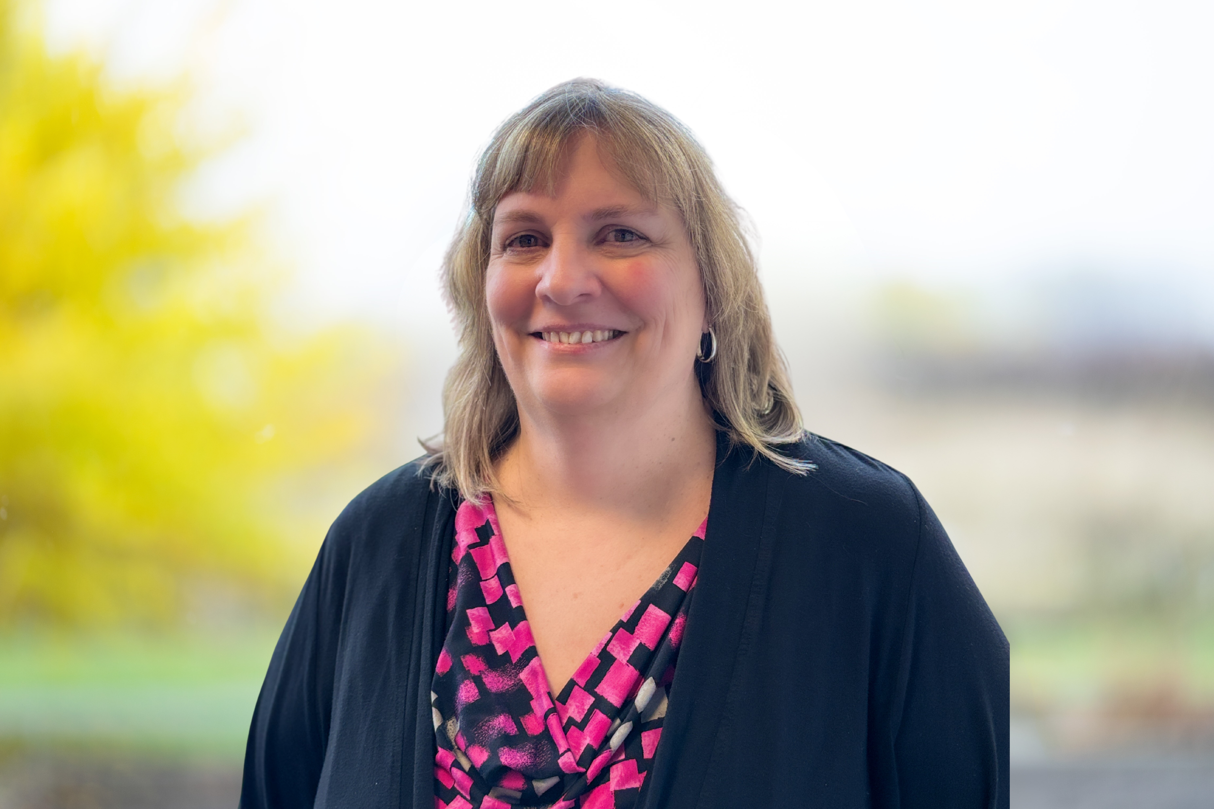 A woman with shoulder-length blonde hair smiling, wearing earrings, a black blazer, and a black and white patterned blouse with red accents, with a blurred outdoor background.