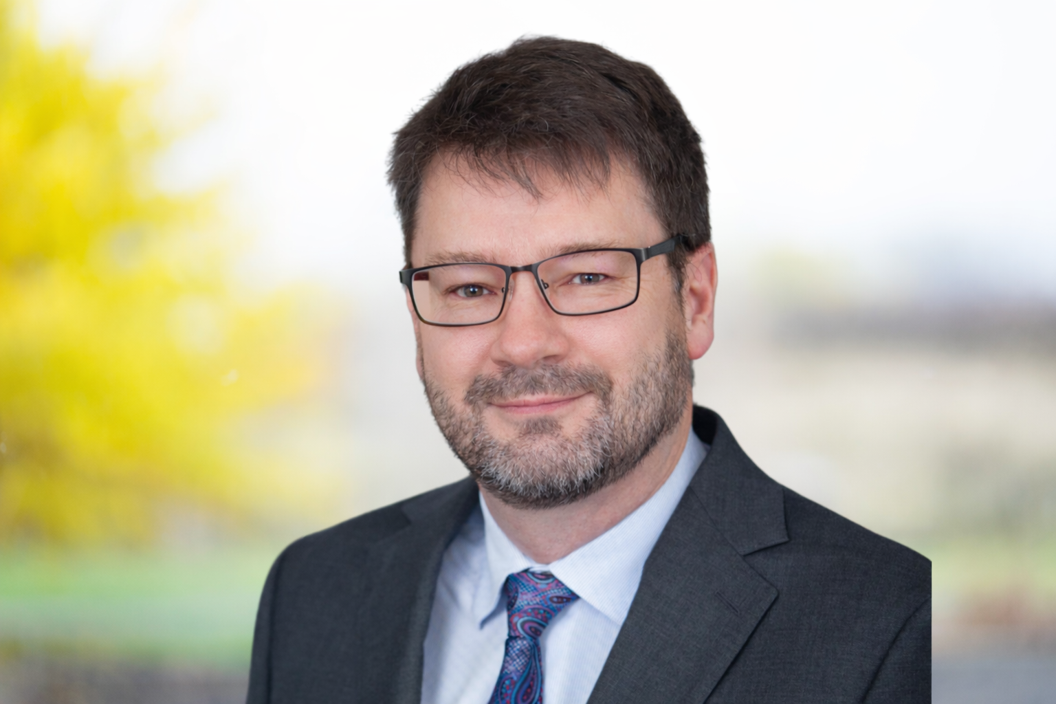 Smiling board member Neil Shay from Caradoc Townsend Mutual in a suit and tie with glasses, standing outdoors with a blurred background of trees.