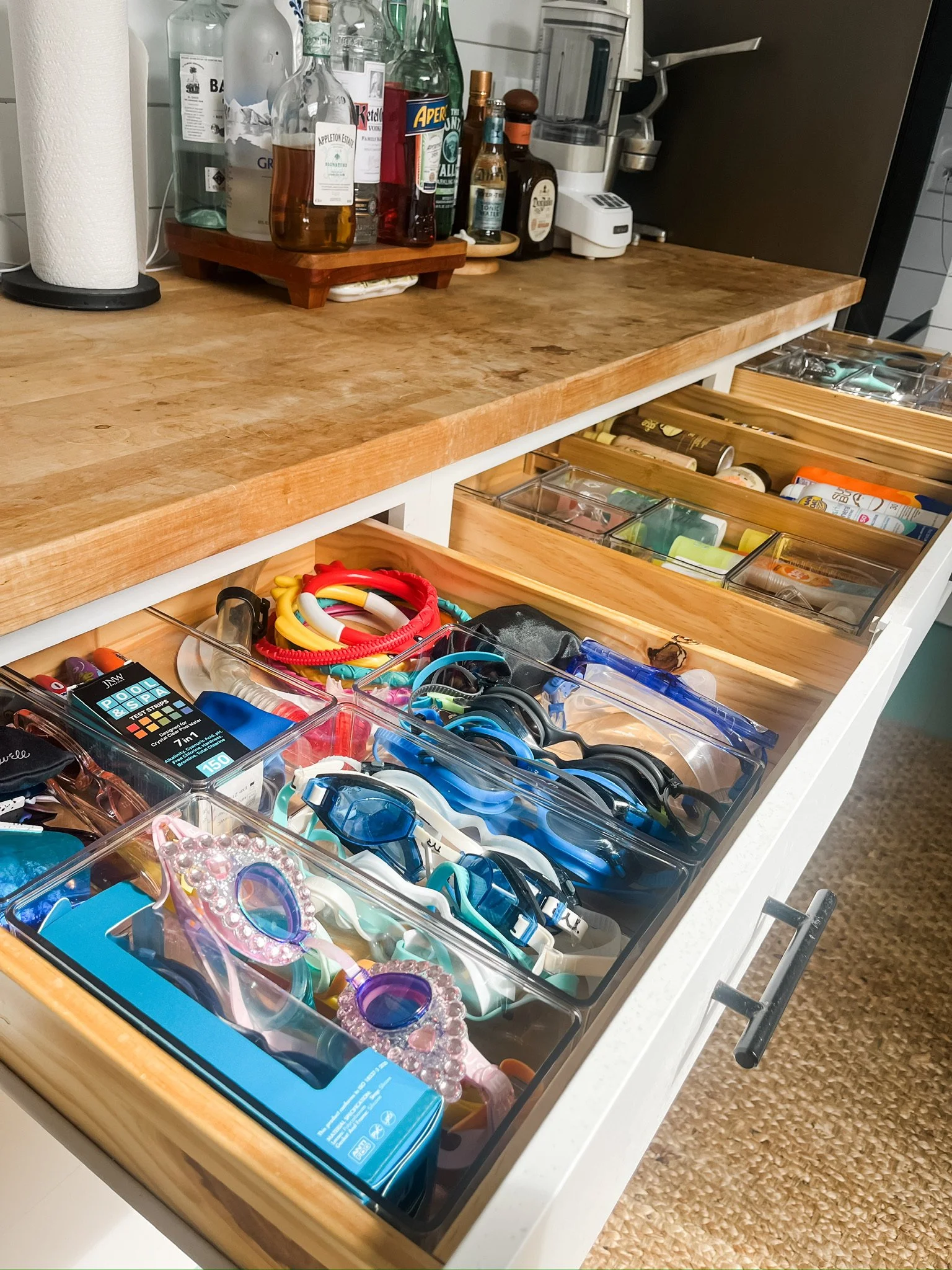 Kitchen drawer organized with sunscreen, sunglasses, bug spray, and other summer essentials stored in clear organizers.