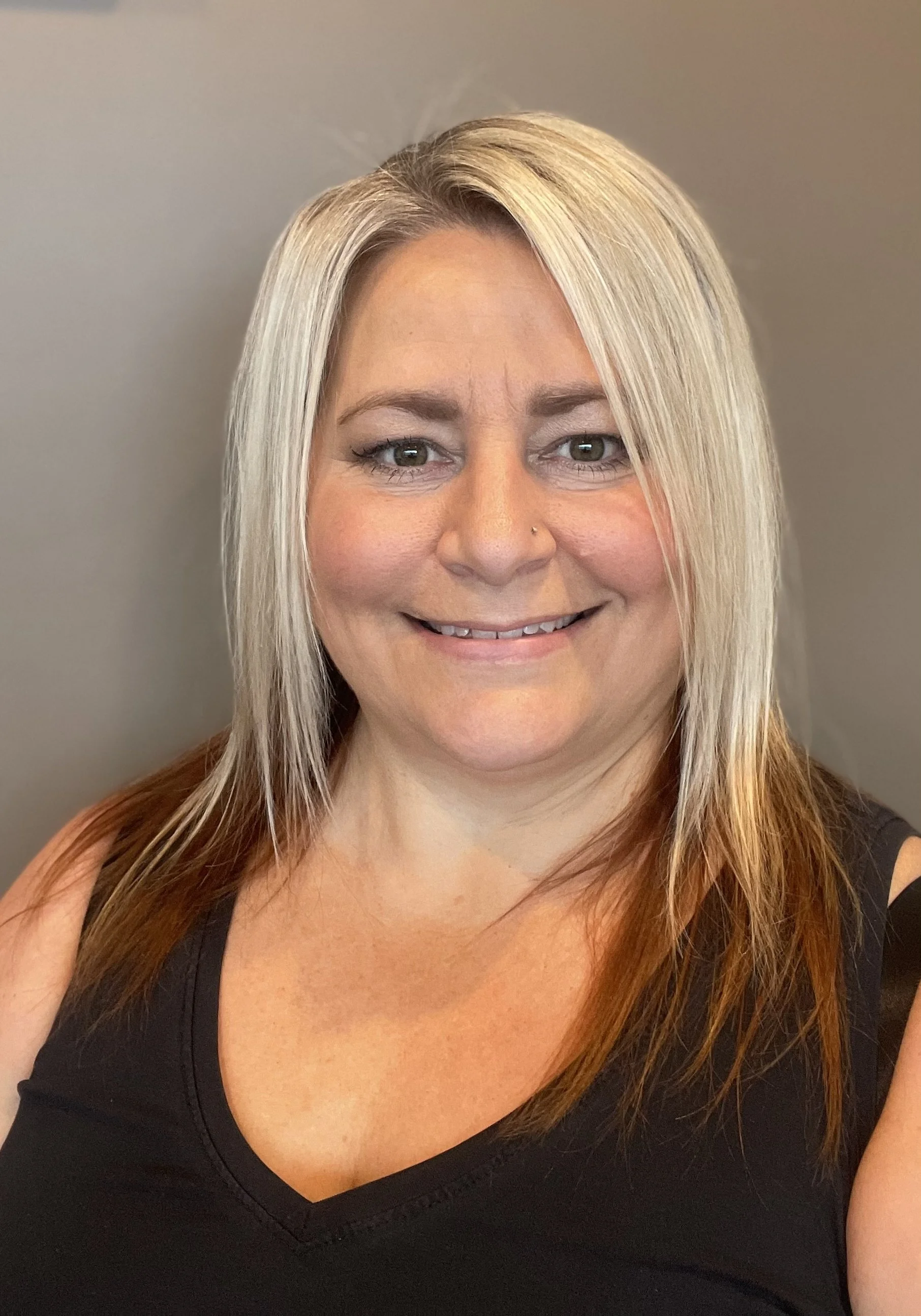 A smiling woman with shoulder-length blond hair, wearing a black sleeveless top, standing against a plain gray wall.