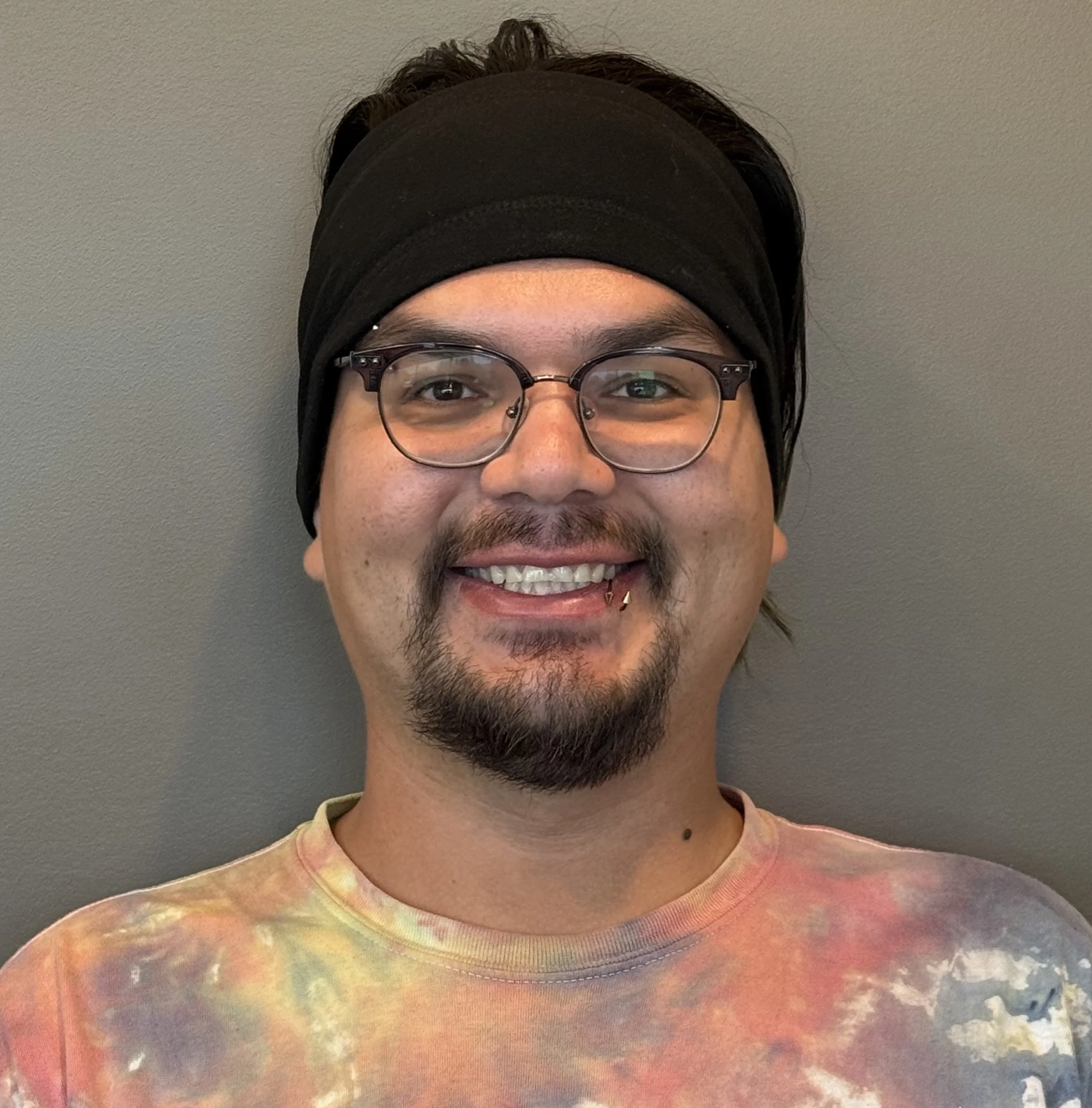 A smiling man with glasses, a goatee, and a lip piercing, wearing a black headband and a tie-dye shirt, standing against a plain wall.
