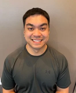 A smiling man with short dark hair wearing a dark athletic shirt, standing in front of a gray wall.