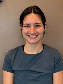 A young woman with dark hair smiling at the camera, wearing a dark gray shirt, against a neutral background.