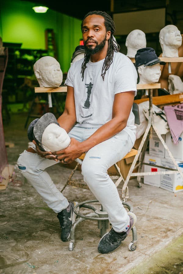 A Black sculptor sits on a stool in a studio space, surrounded by plaster heads.