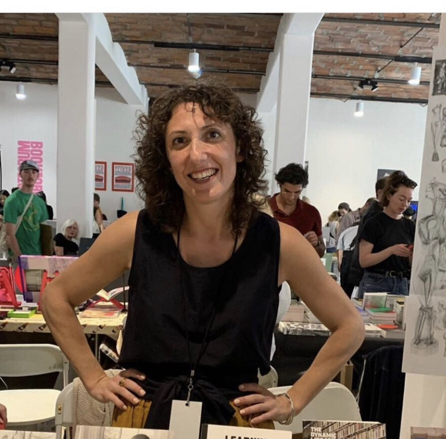 Image: Julia Klein, wearing a black tank top and smiling for the camera, poses at her booth at the New York Art Book Fair. Photo by Lisa Pearson.