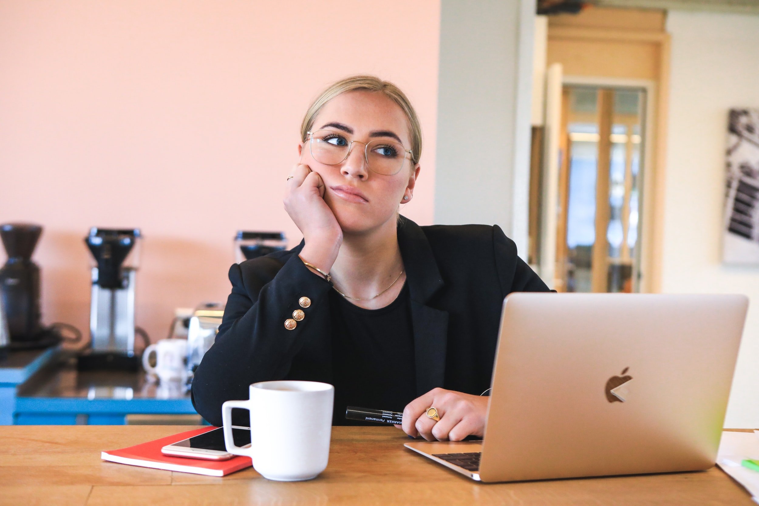 A young white woman sits behind a desk with her head in her palm. She appears to be a dejected job seeker wearing a suit and sitting behind a computer.