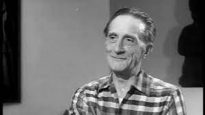 Marcel Duchamp at approximately age 70 wearing a plaid shirt and smiling in a televised interview.