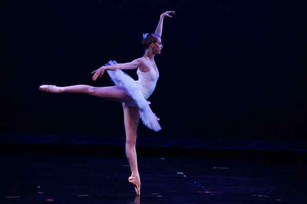 A ballerina dances on a stage, her body upright with her left arm overhead and right arm extended back, toward her outstretched right foot.