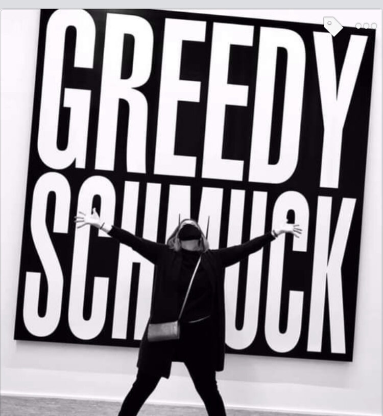 A black-and-white image of a woman in black winter clothing in front of a Barbara Kruger artwork that says "Greedy Schmuck"