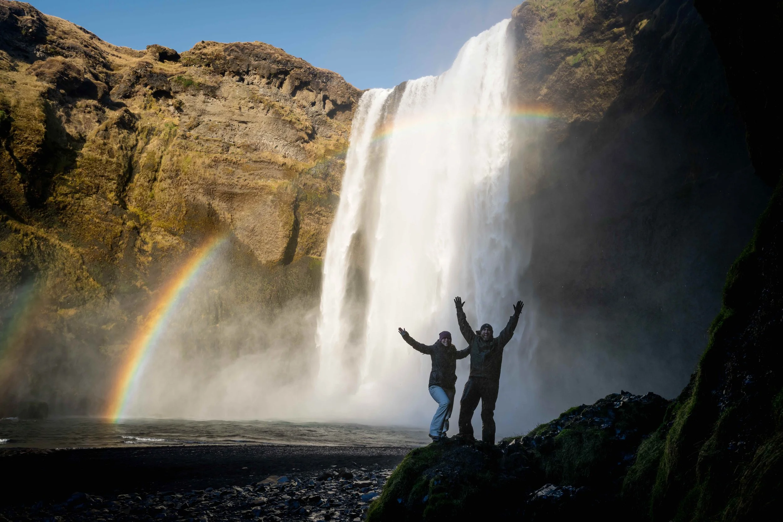Two people standing with arms raised in front of a large waterfall with rainbow and mist