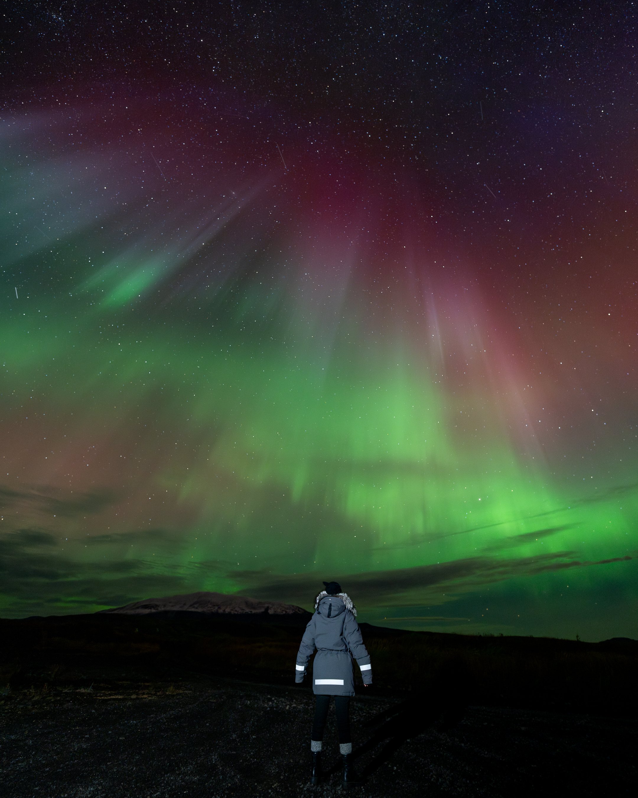 A person dressed in winter clothing standing in darkness and looking up at the night sky illuminated by the Northern Lights and stars.