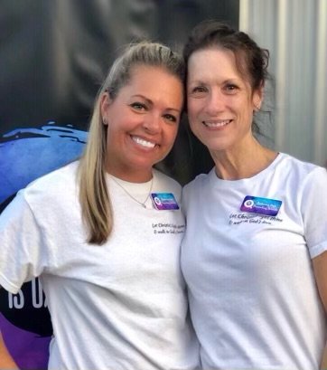 Two smiling women in matching white t-shirts, each wearing a name badge, standing side by side in front of a dark background with blue and purple elements.
