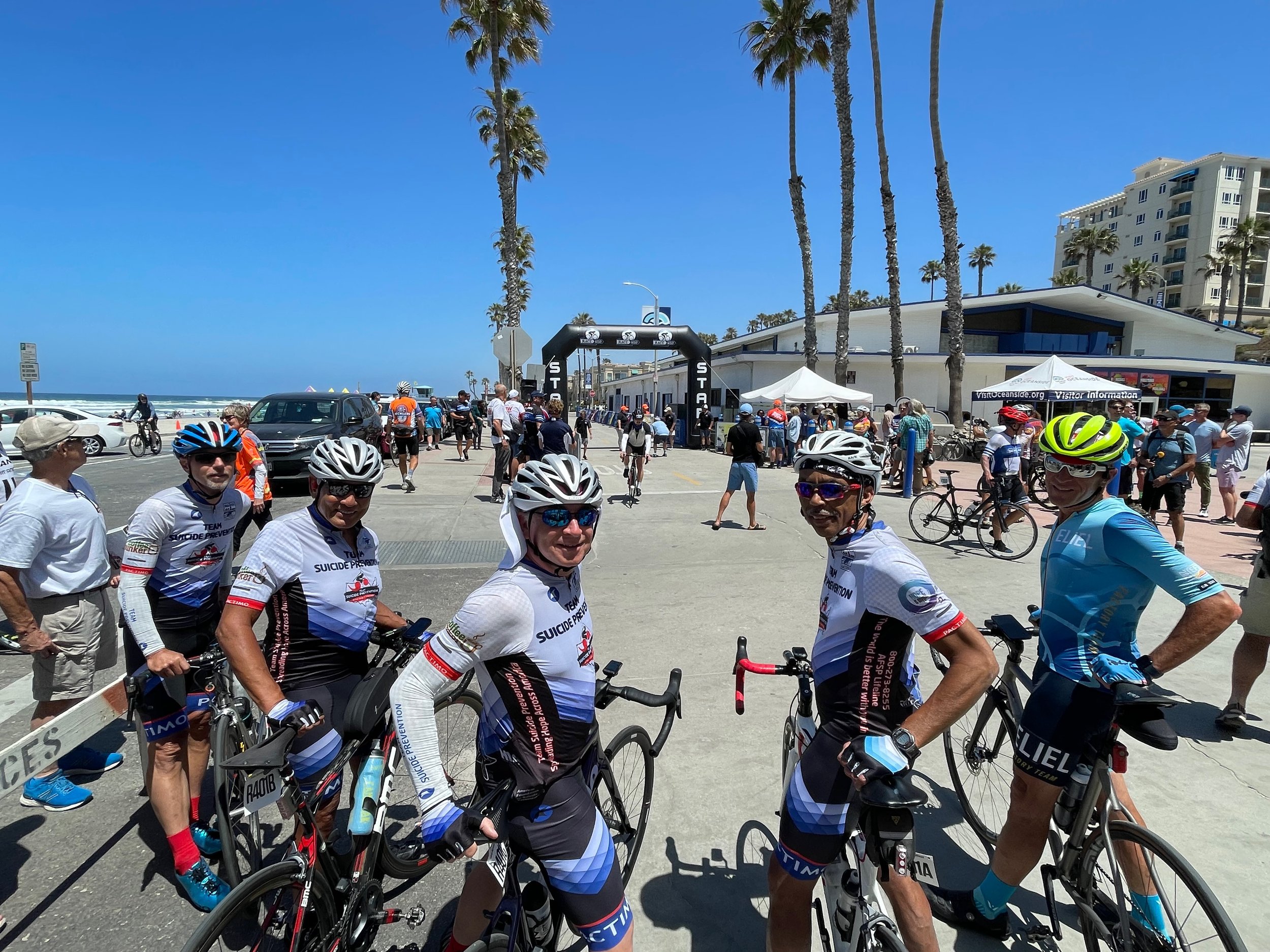 Cyclists in helmets and jerseys smiling at a cycling event near a start line with palm trees and ocean in the background.
