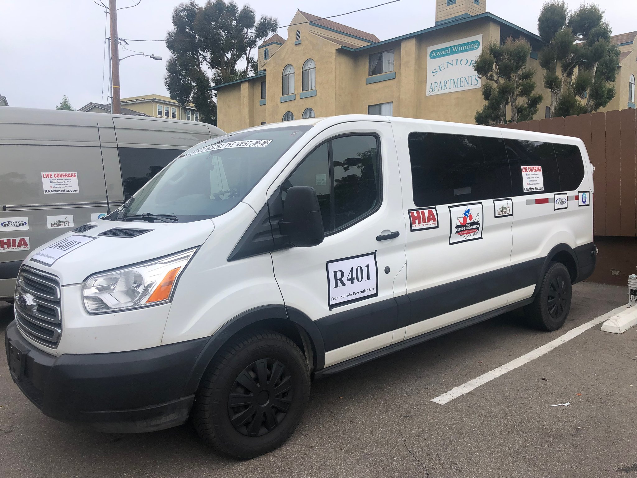 White Ford van labeled "R401 Team Suicide Prevention One" parked beside buildings labeled "Senior Apartments," featuring various logos and stickers.