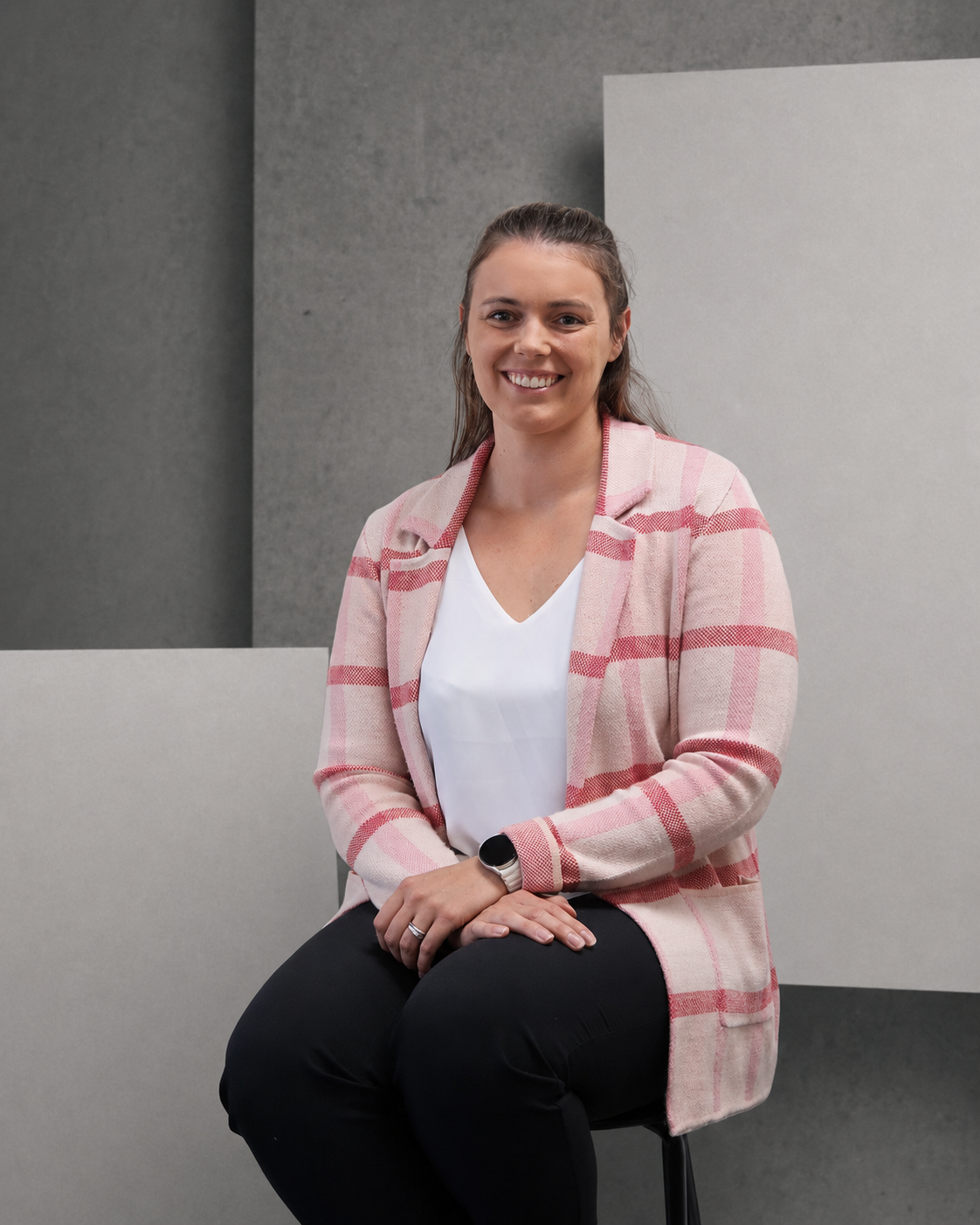 A woman with brown hair, wearing a pink jacket, sitting on a black stool in front of a minimalist background with gray and white walls. She is smiling and looking to the camera.
