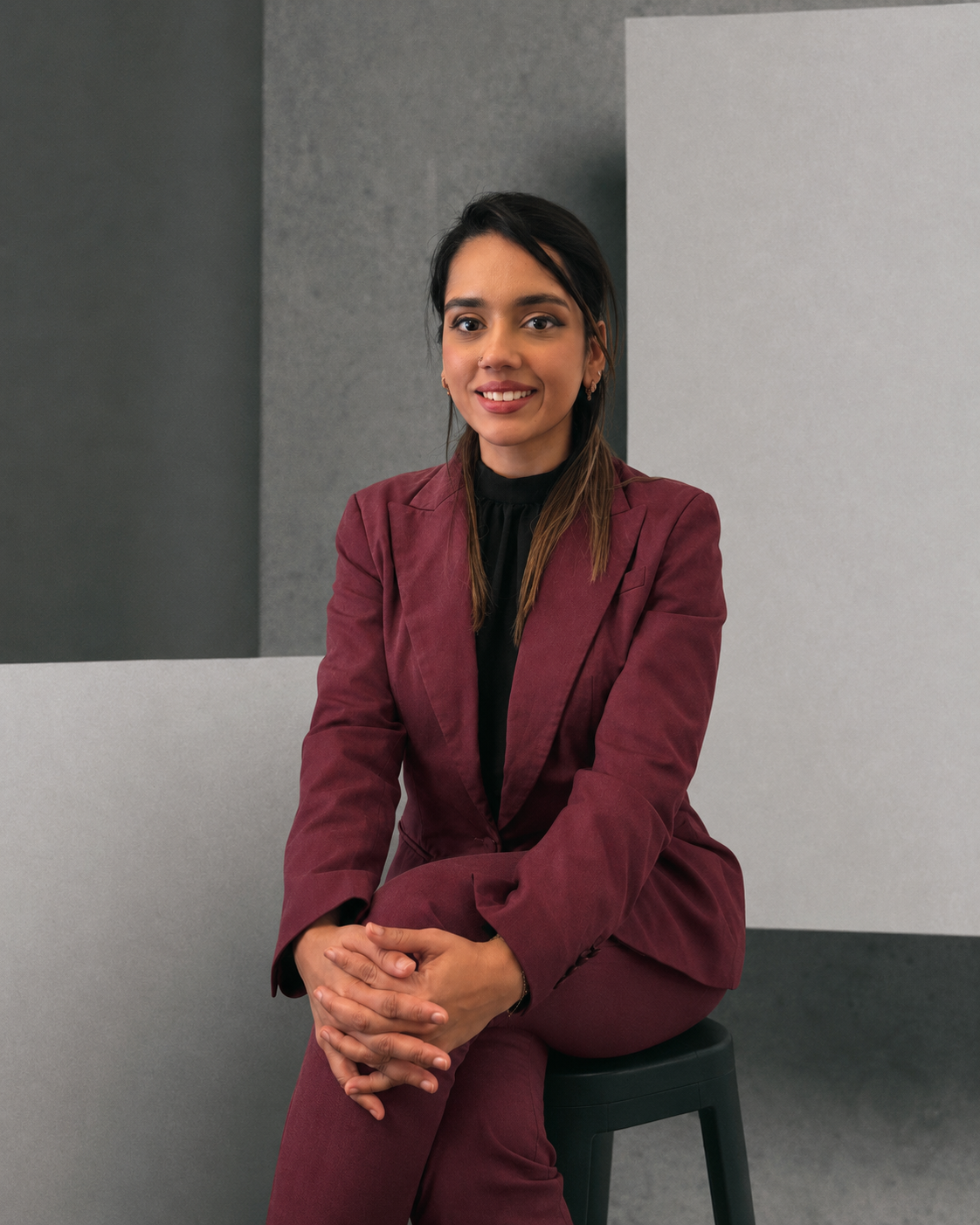 A woman with dark brown hair, wearing a red jacket, sitting on a black stool in front of a minimalist background with gray and white walls. She is smiling and looking to the camera.