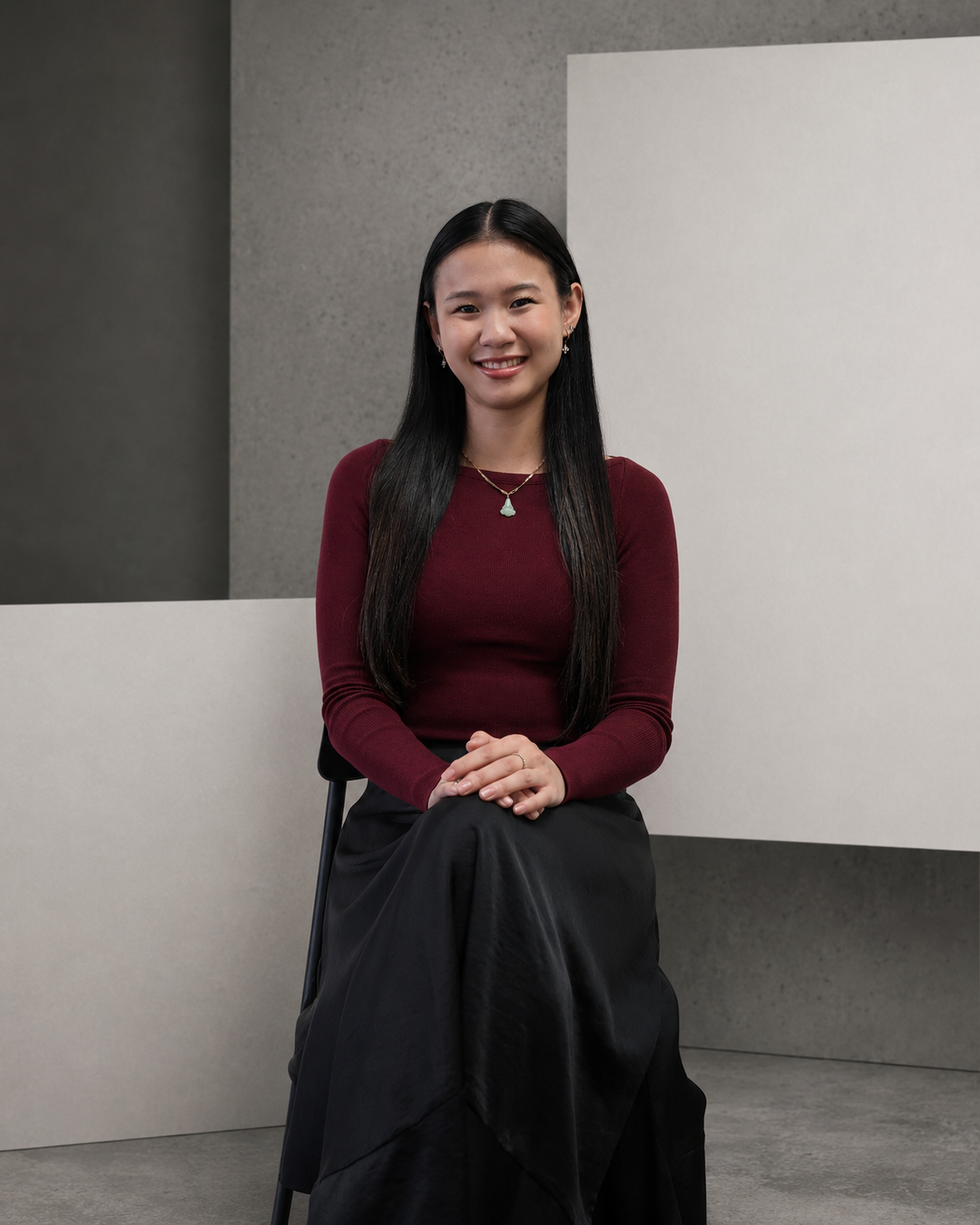 A woman with black hair, wearing a red top, sitting on a black stool in front of a minimalist background with gray and white walls. She is smiling and looking to the camera.