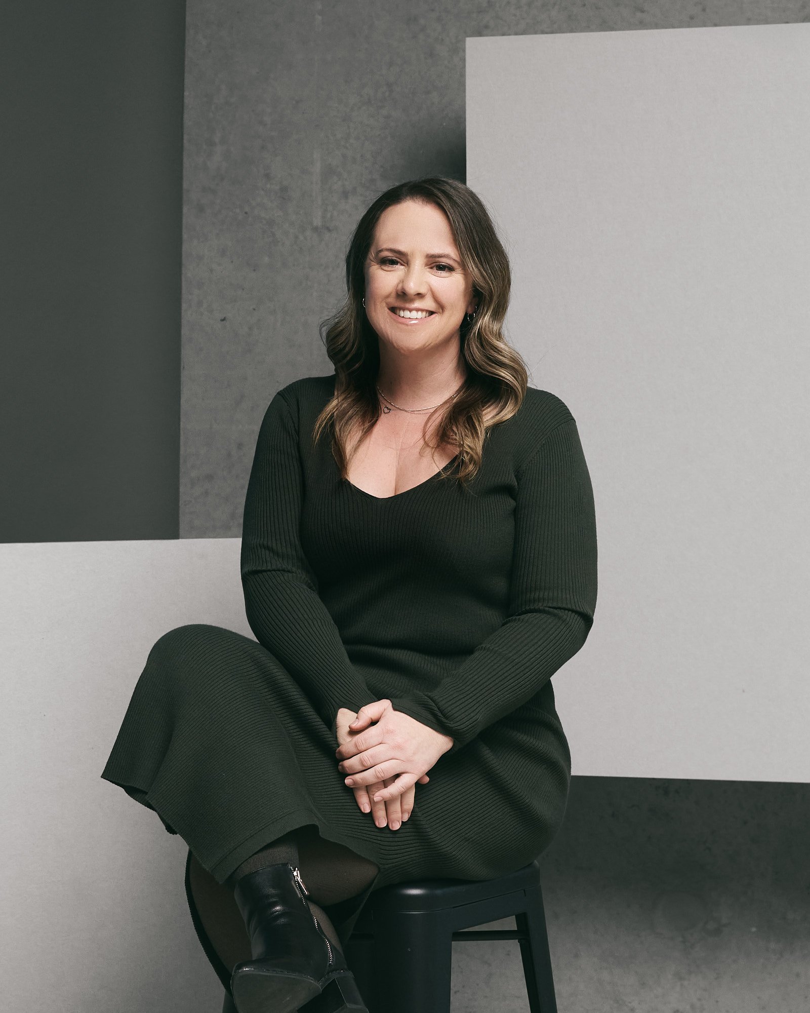 A woman with brown hair, wearing a green dress, sitting on a black stool in front of a minimalist background with gray and white walls. She is smiling and looking to the camera.