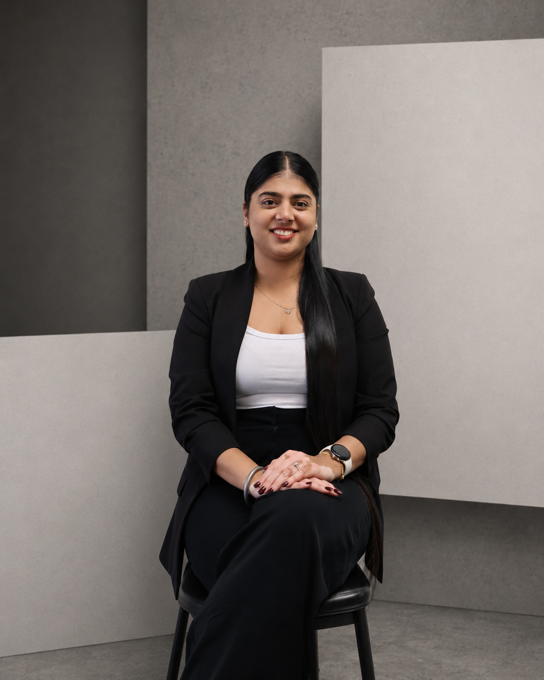 A woman with black hair, wearing a black jacket, sitting on a black stool in front of a minimalist background with gray and white walls. She is smiling and looking to the camera.