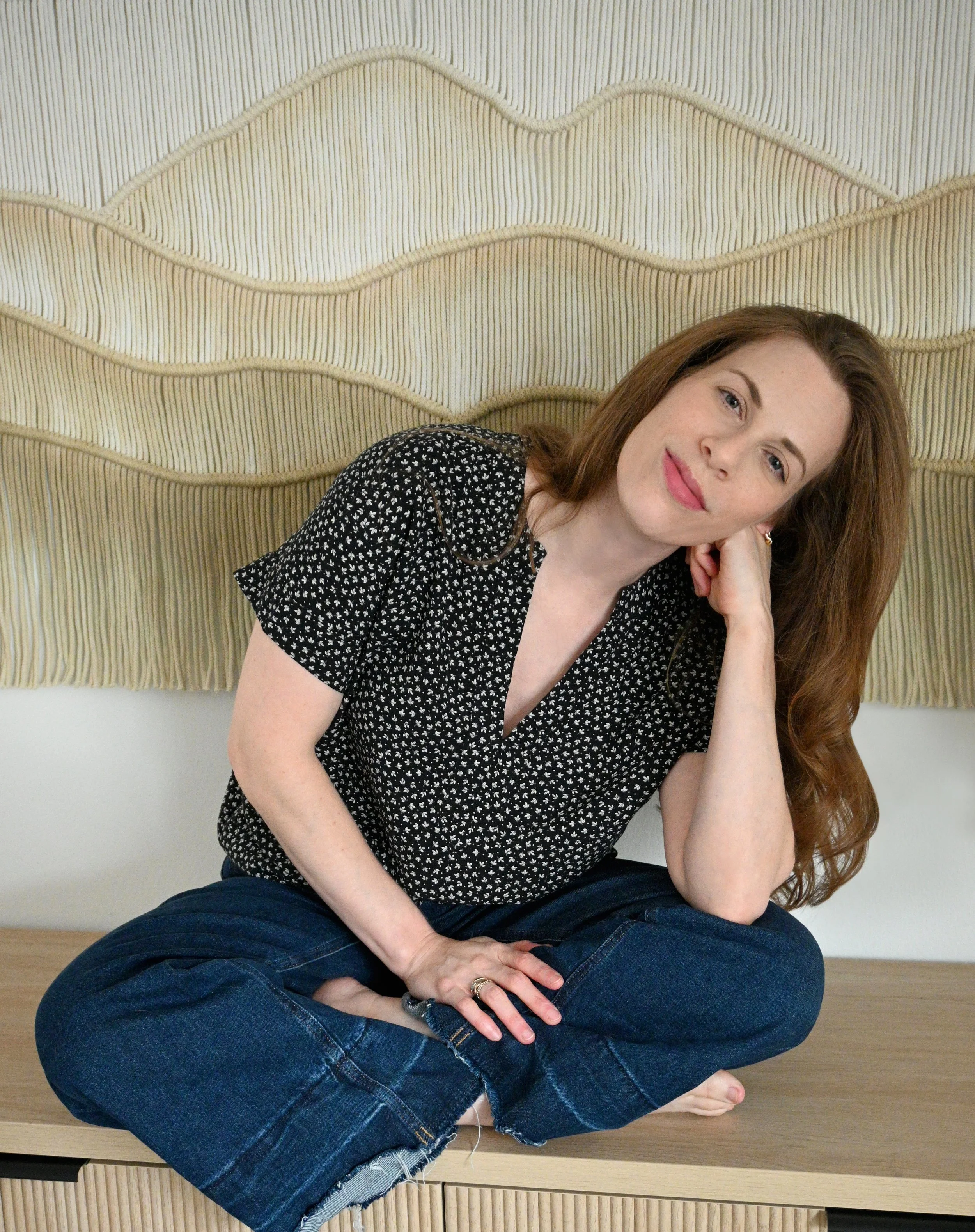 Samantha Paige Rosen sitting cross-legged against a macreme mountain backdrop wearing jeans and a black and white patterend shirt with blue eyes, reddish hair, closed-mouth smile