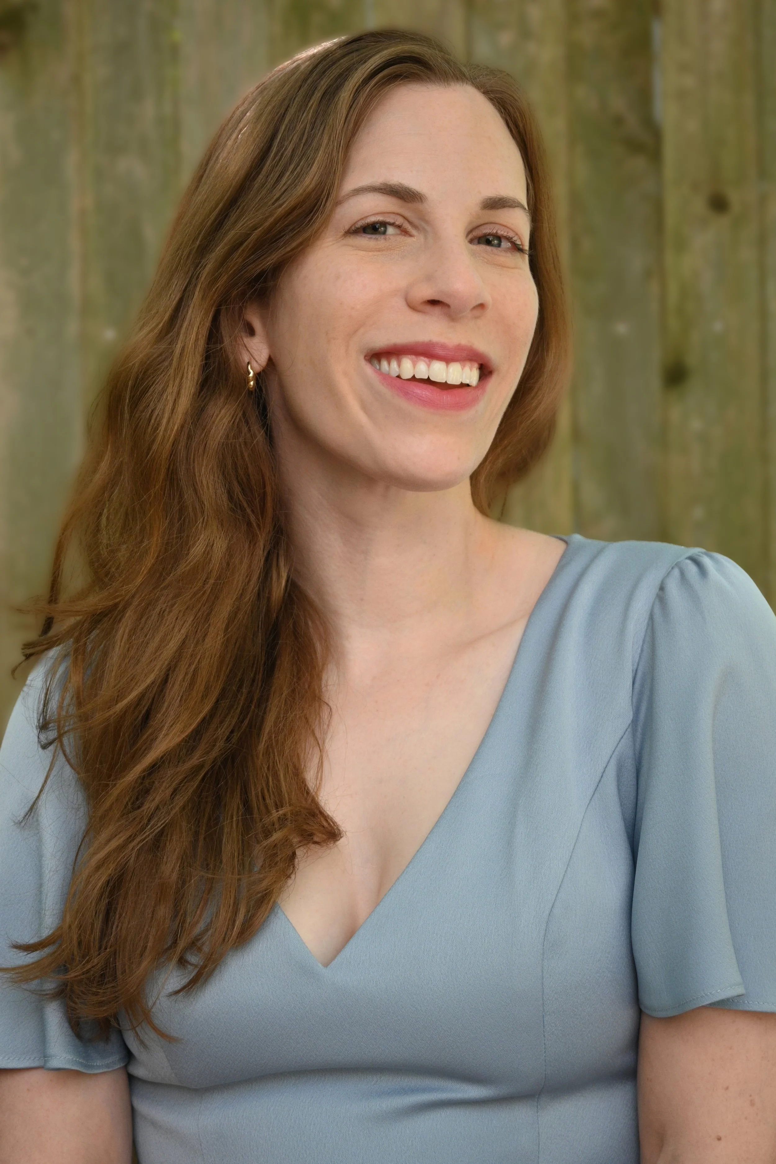 Samantha Paige Rosen smiling in front of a fence and looking over her left shoulder. She's wearing a light blue shirt and has blue eyes and reddish hair.
