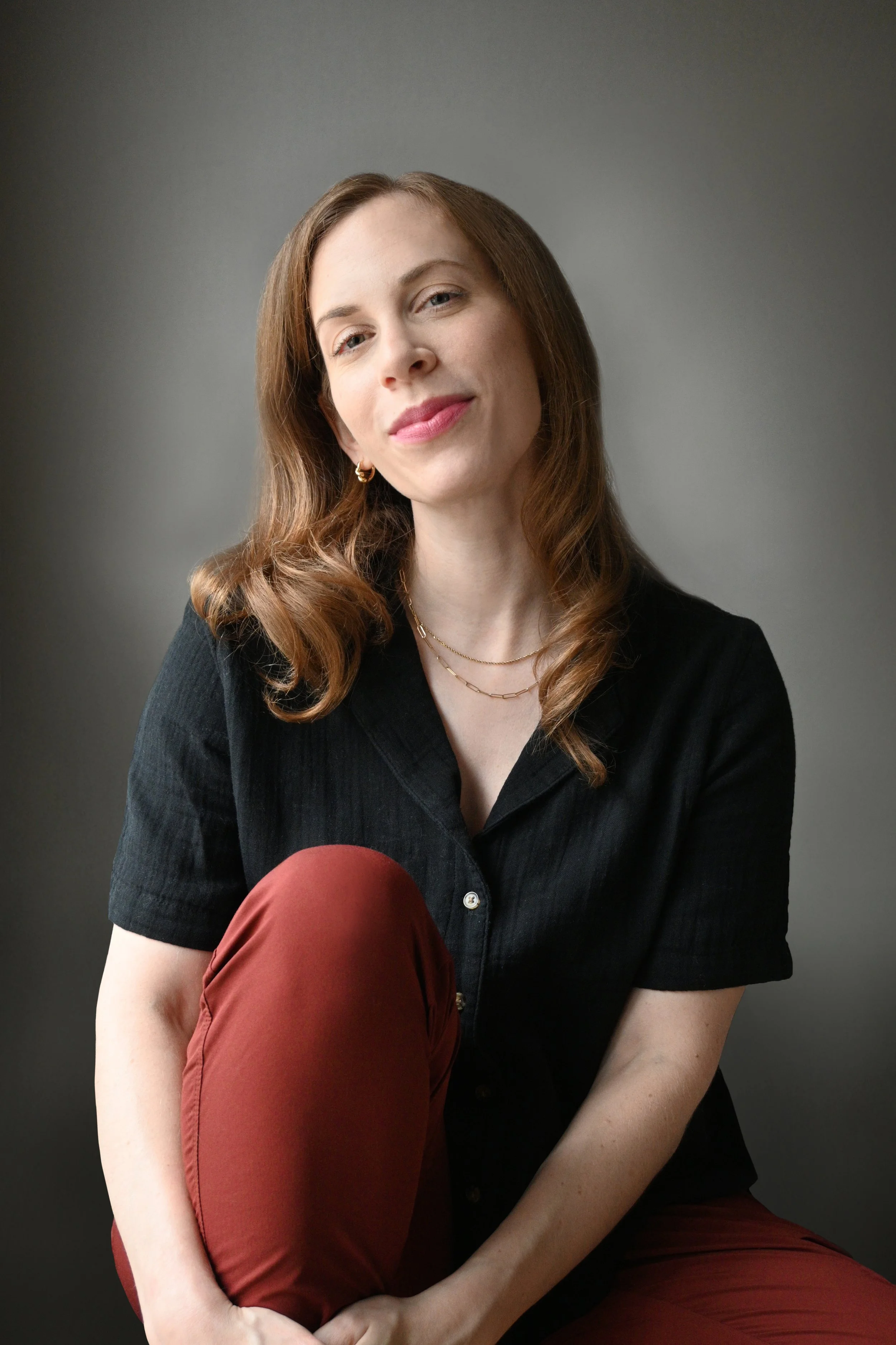 Reddish-blond, blue-eyed woman, Samantha Paige Rosen, in a black button-down shirt orange pants, sitting in a chair with one knee up