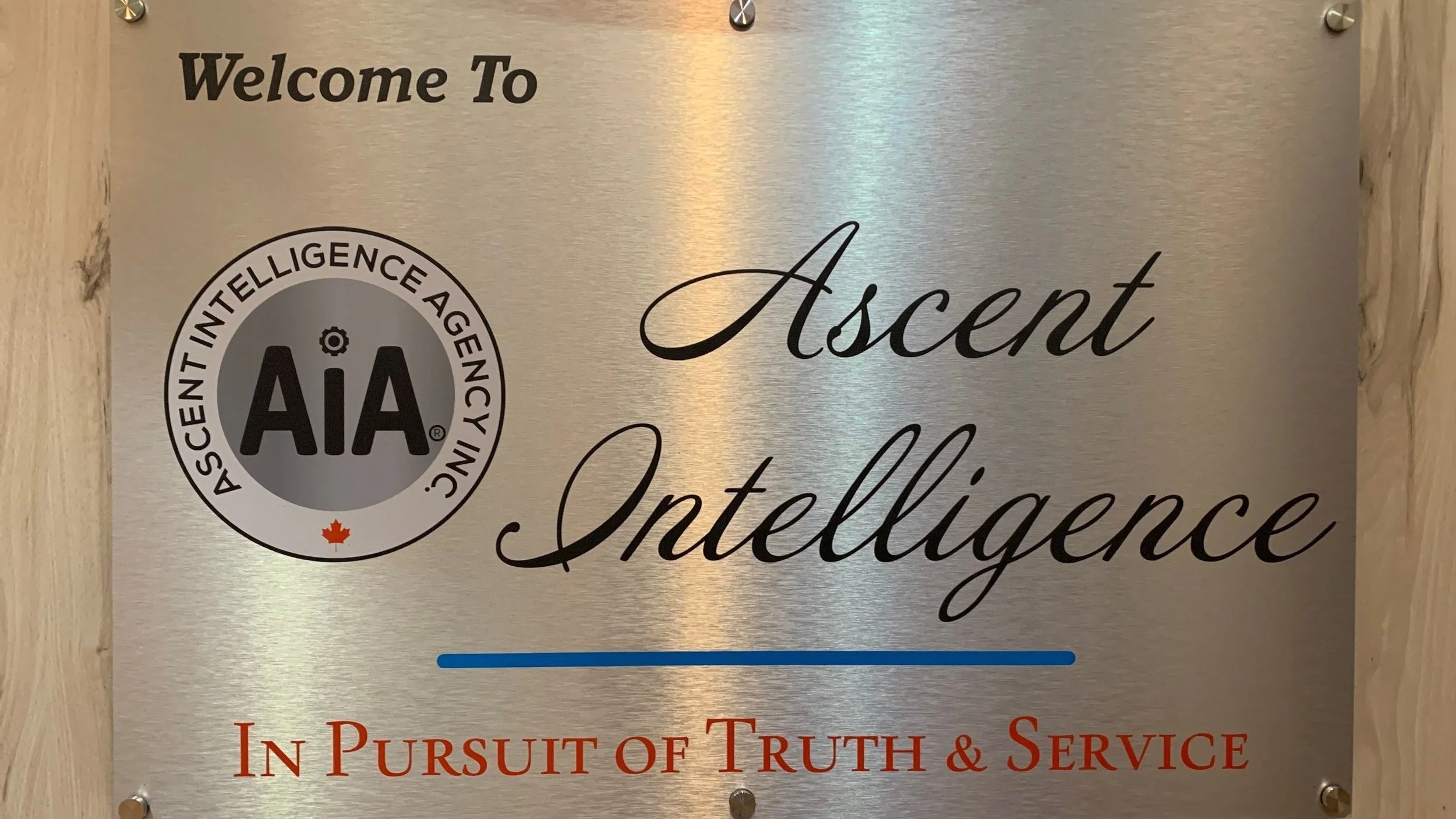 Metallic sign reading "Welcome To Ascent Intelligence" with the logo of Ascent Intelligence Agency Inc. (AIA) on the left, featuring a small maple leaf under the logo. The bottom text says "In Pursuit of Truth & Service."