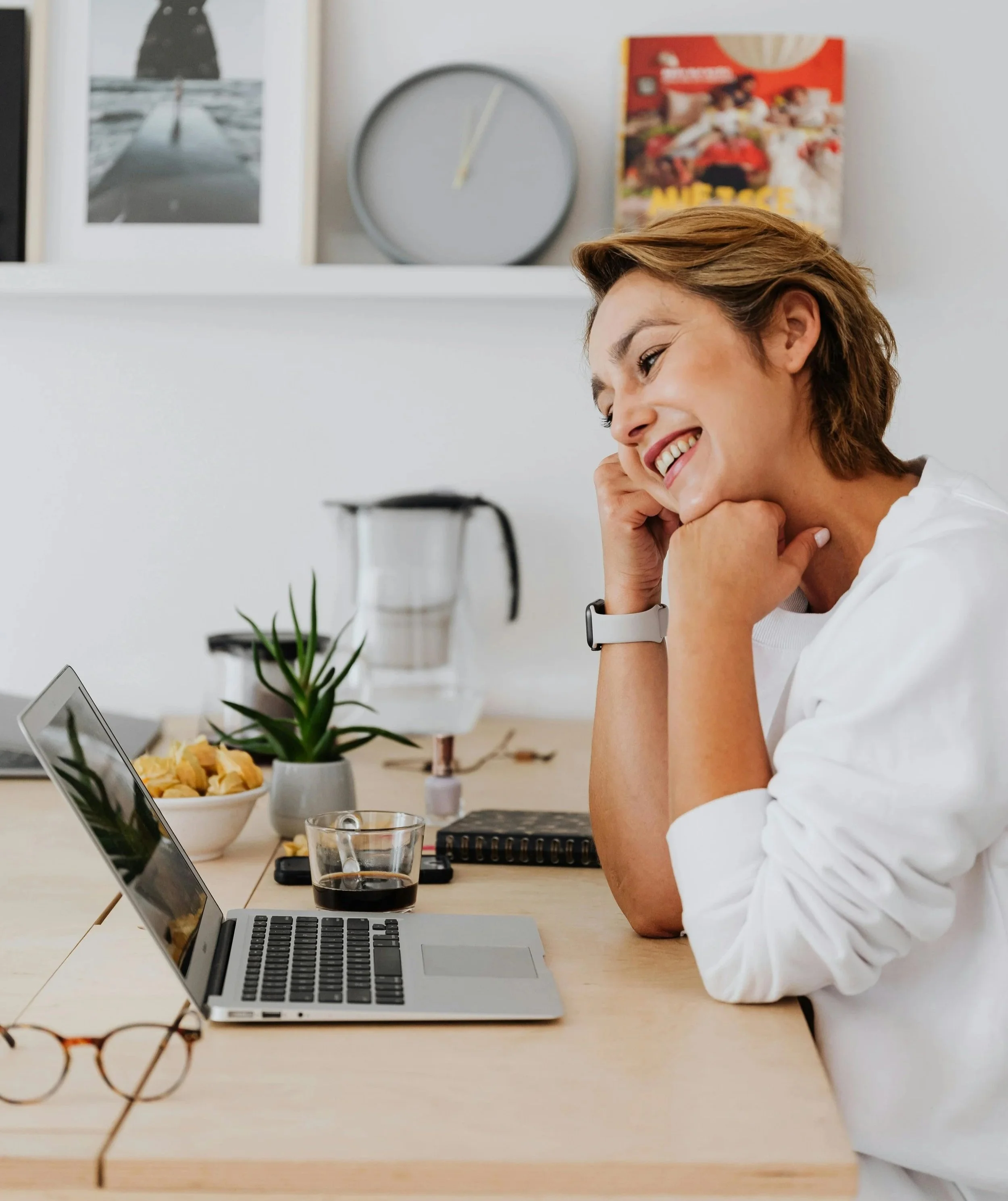 A woman sitting at a wooden desk, smiling while looking at her laptop screen, with a glass of coffee, a potted plant, and a bowl of snacks on the desk, in a cozy home office setting.