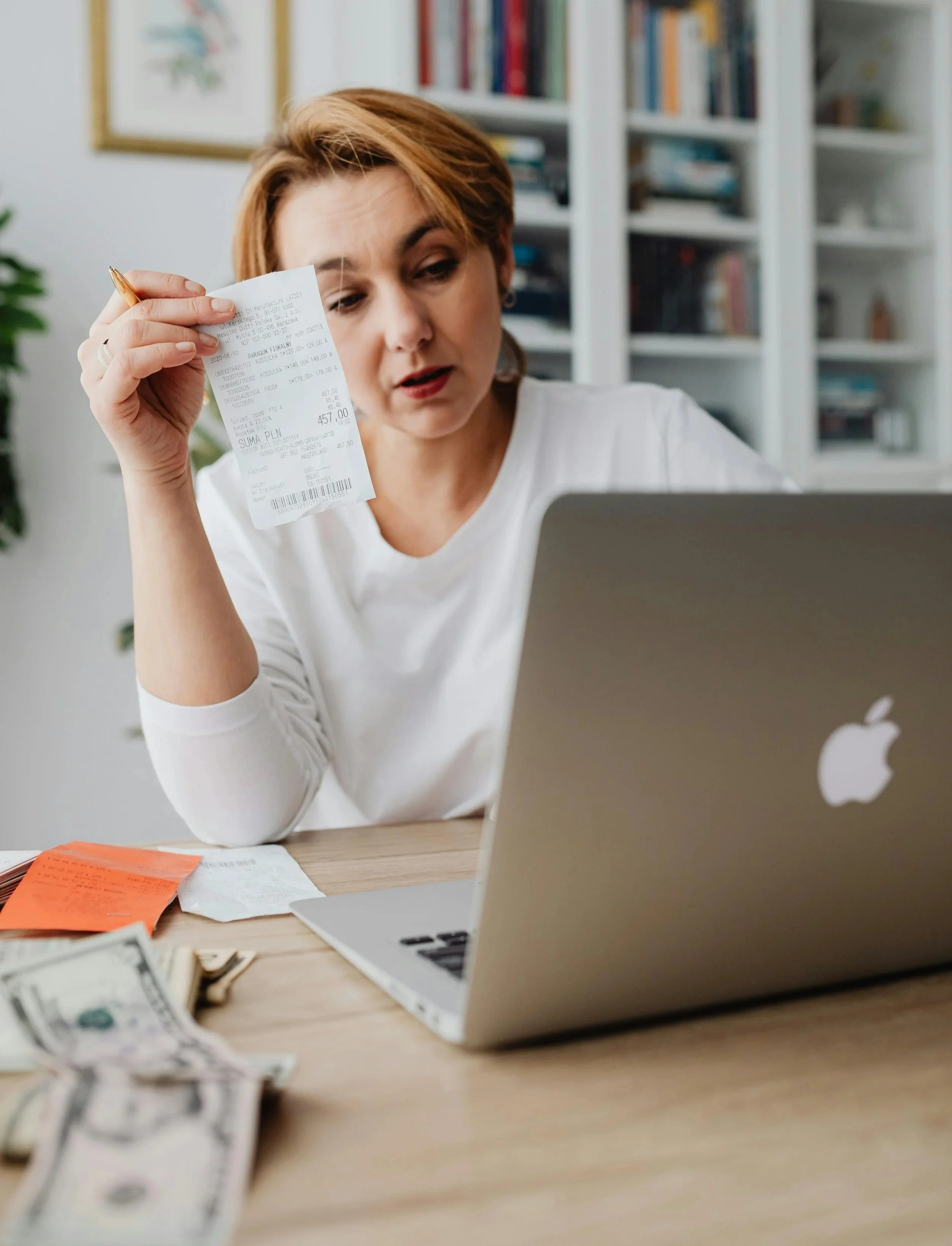 A woman looks at a receipt with a confused expression while sitting at a desk with a MacBook laptop, cash, and receipts.