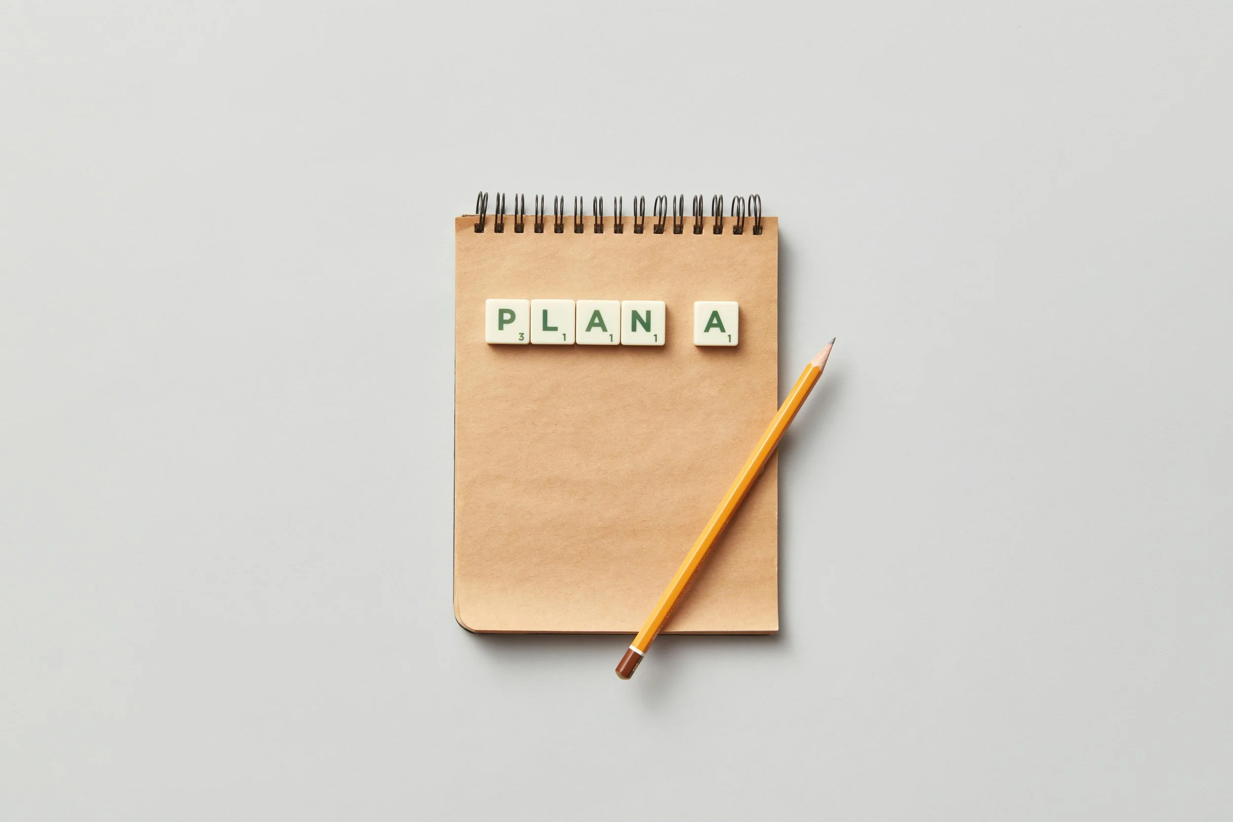 A notebook with Scrabble tiles spelling 'PLANA' on top, accompanied by a yellow pencil on a white background.