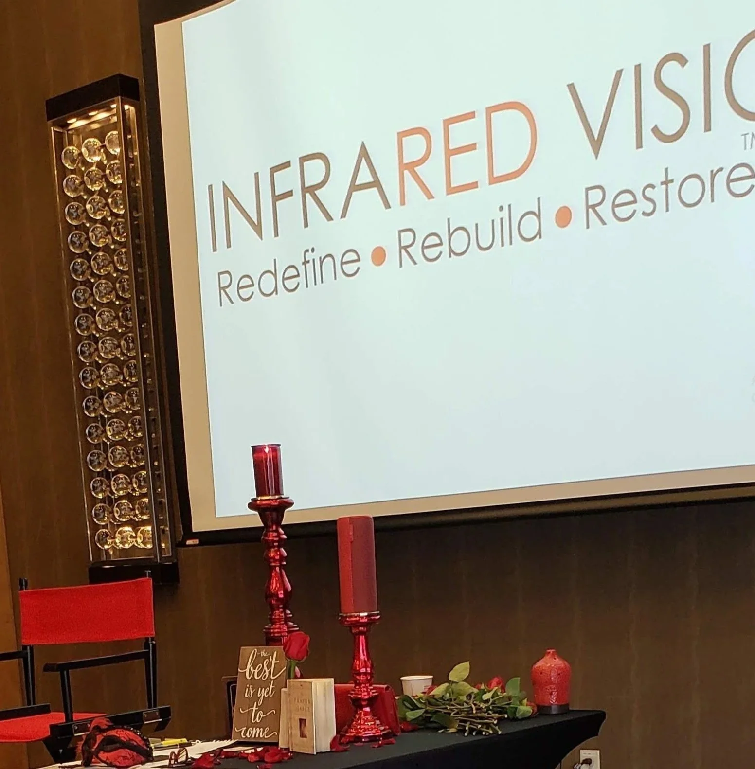 Projection screen displaying 'Infrared Vision' with the words 'Redefine, Rebuild, Restore'. A table in front has red candles, a red decorative vase, greenery, and a sign that reads 'The best is yet to come'.