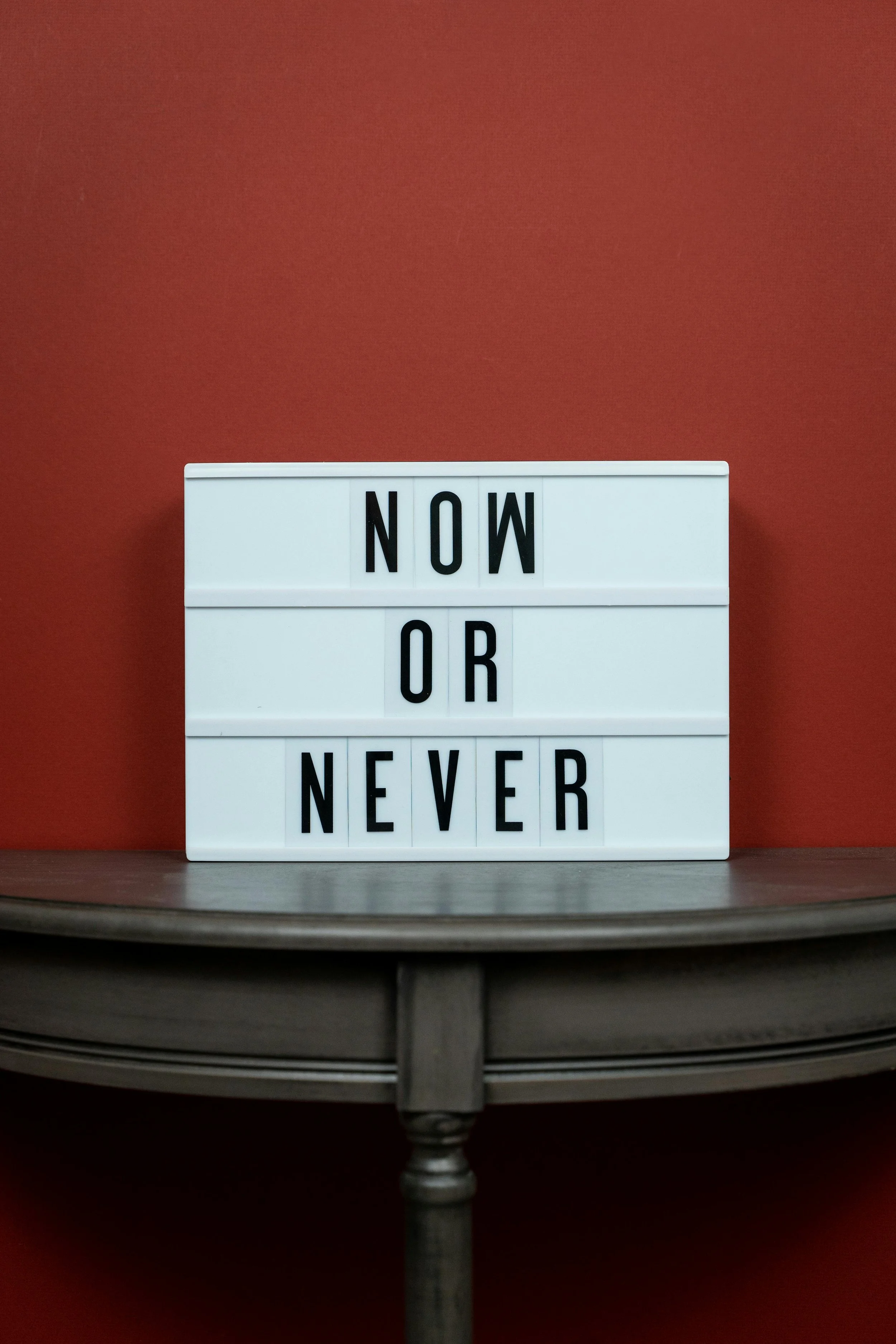 A lightbox sign displaying the words 'NOW OR NEVER' against a red wall, placed on a wooden table.