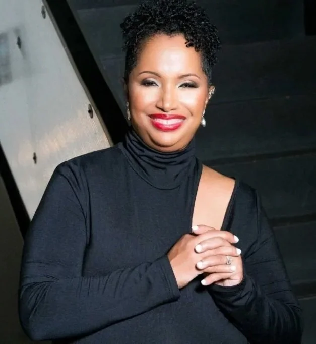 A woman with short curly black hair, red lipstick, and earrings, wearing a black top with a slit, smiling and clasping her hands together.