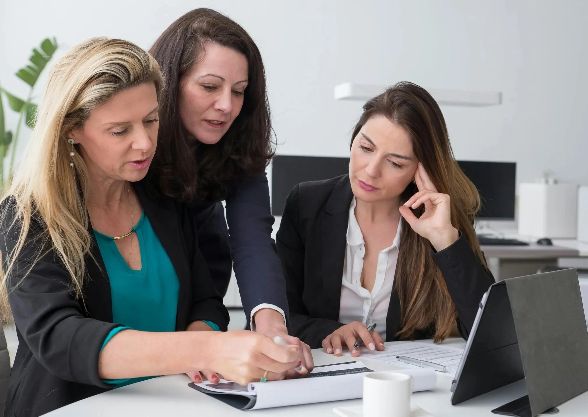 Three women in business attire looking at documents and a tablet in an office.