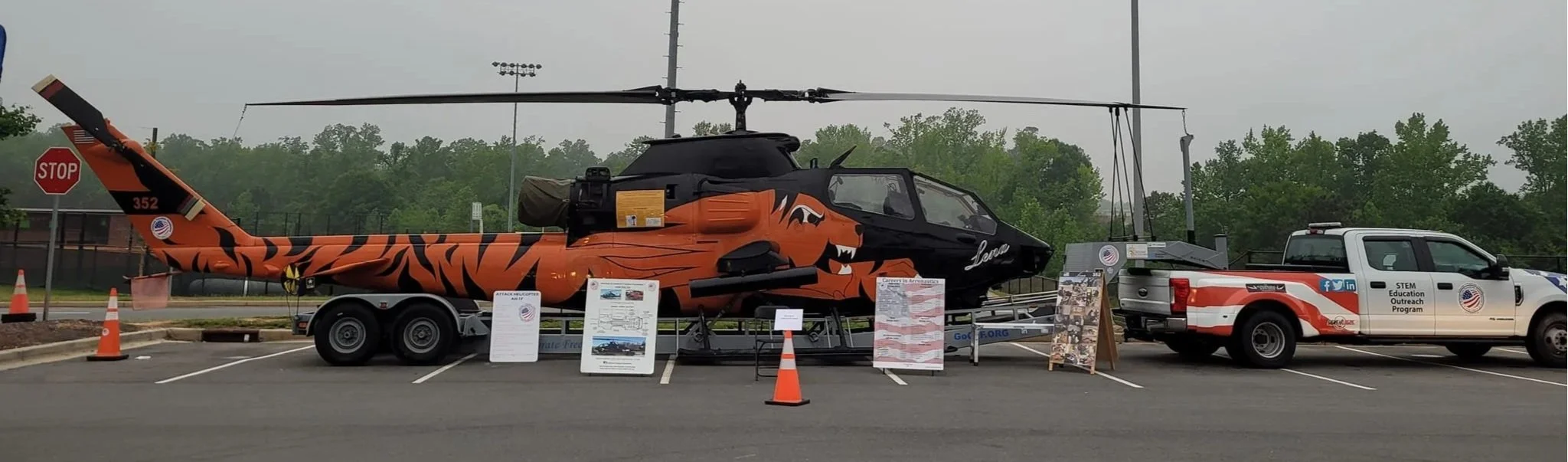 Display of a helicopter with a tiger stripe design on a trailer, set up in a parking lot with informational signs and orange traffic cones, and a white utility truck labeled 'STEM Education Outreach Program' parked nearby.