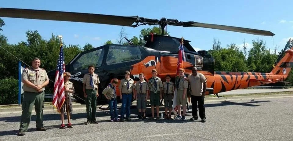 Group of Boy Scout and adult leaders standing in front of a military helicopter, with American flags displayed.