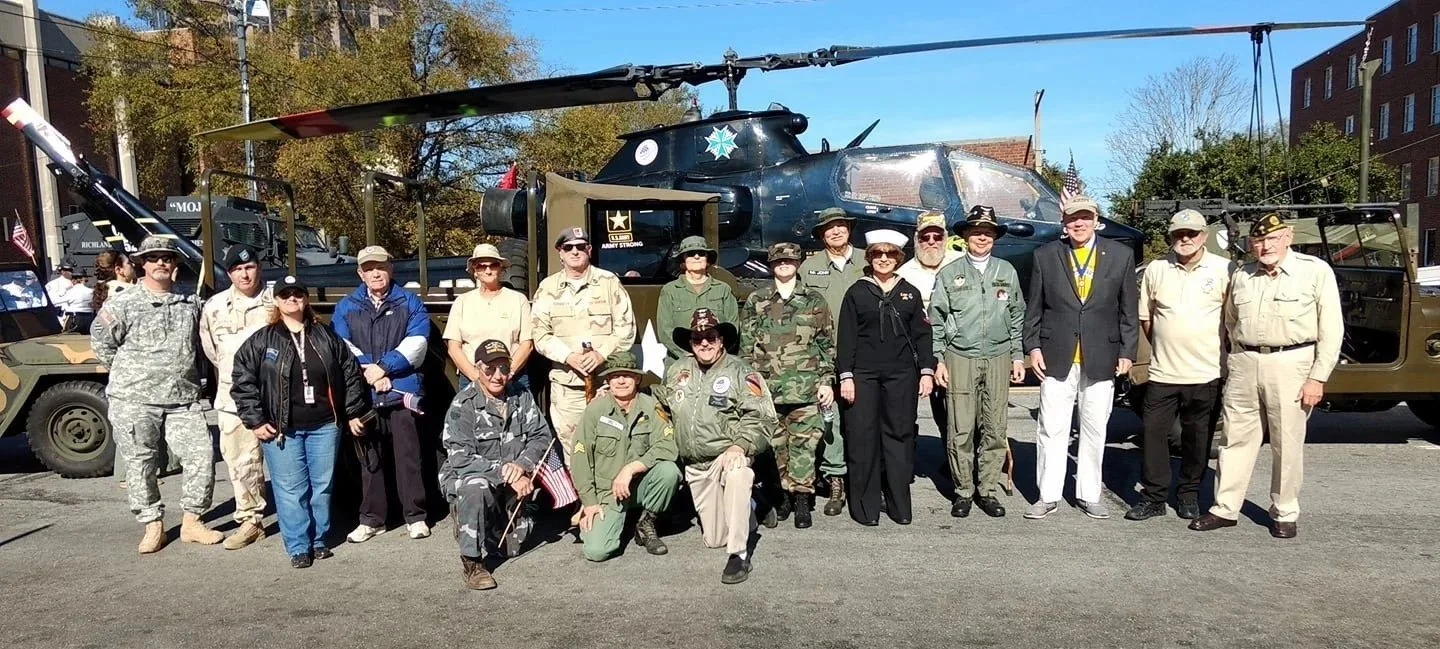Group of people dressed in military and civilian clothing standing in front of a large military helicopter and vintage military vehicles, outdoors on a sunny day.