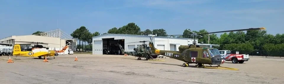 Military vehicles, including helicopters and a jet, parked on an airfield with hangars in the background on a clear day.