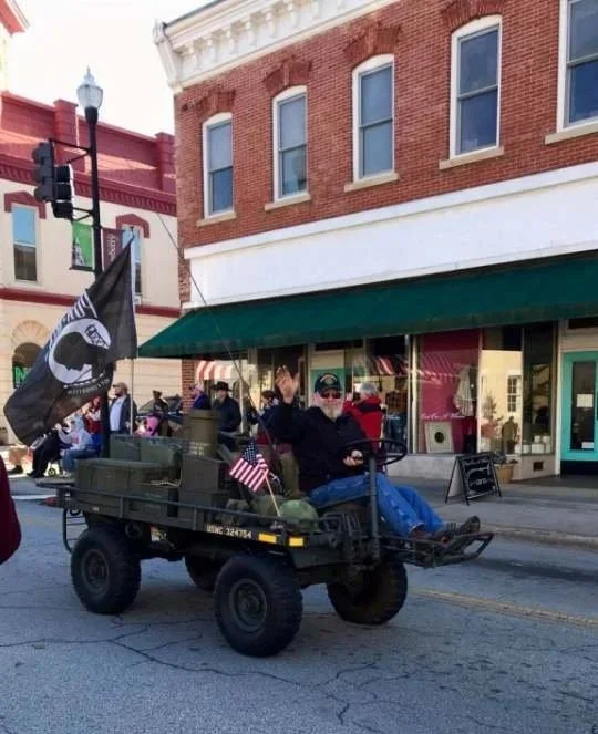 A person riding in a small, military-style flatbed vehicle decorated with American flags and a pirate flag during a parade or street event, with spectators in the background.