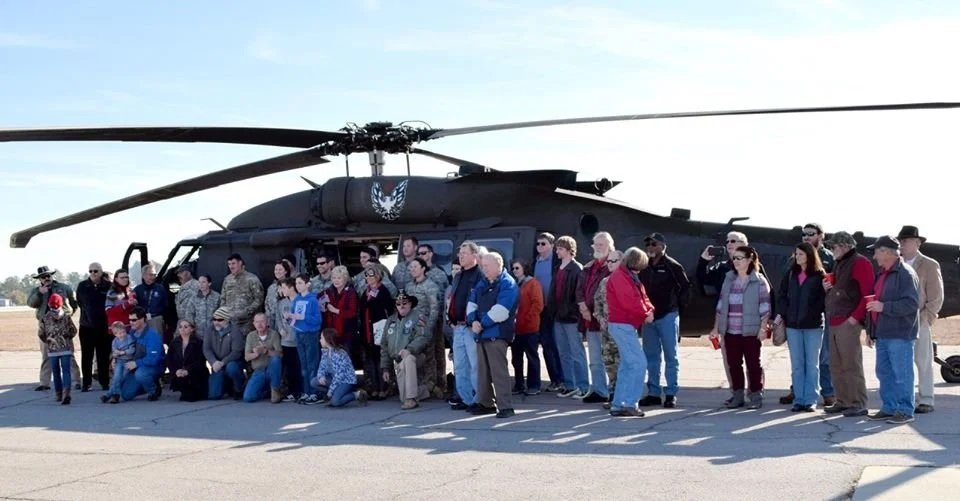 Group of people gathered around a military helicopter on a tarmac, with some kneeling and others standing, during daytime.