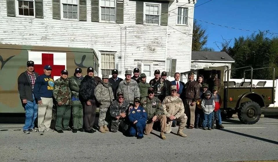 Group of veterans and children standing in front of military and ambulance vehicles on a street.