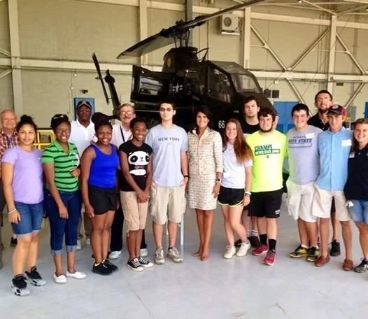 Group of students and a woman standing inside an aircraft hangar, posing in front of a military helicopter.