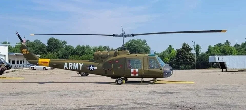 Military helicopter painted in army colors with a red cross on the side, parked on an airport tarmac under a partly cloudy sky.