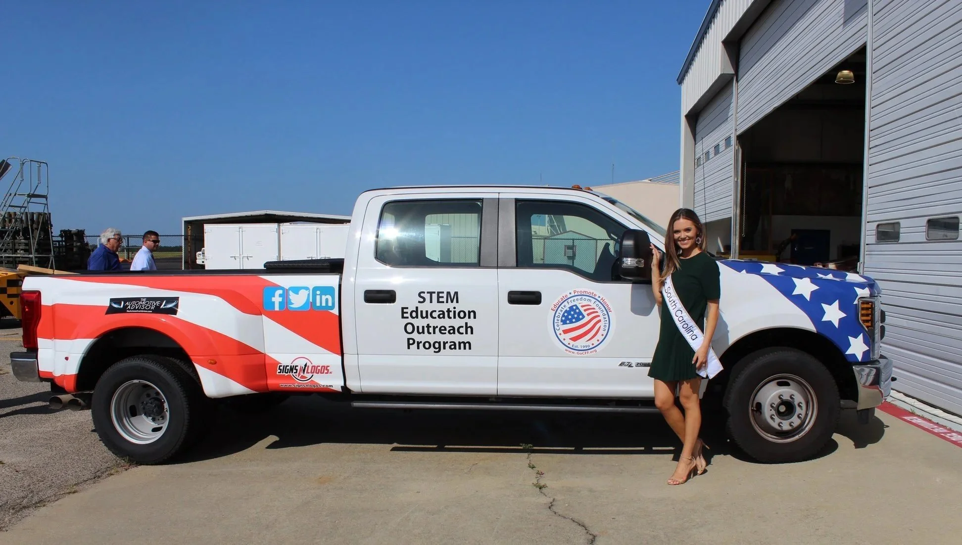 A woman standing next to a truck with patriotic red, white, and blue decals for a STEM education outreach program, smiling and wearing a sash that says 'South Carolina'.