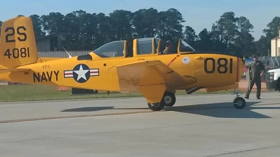 A yellow vintage aircraft parked on a tarmac with two people nearby, one inspecting the aircraft and the other walking.