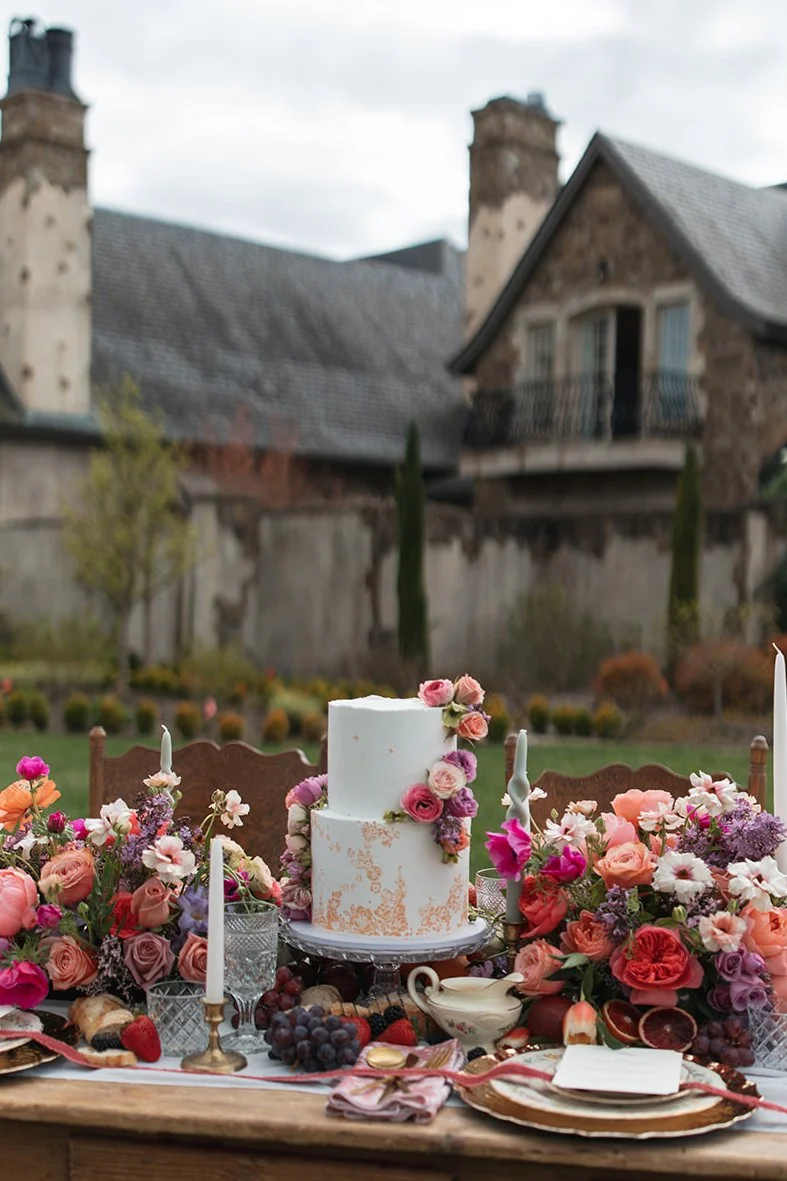   Elegant Portland Oregon wedding cake displayed on a styled dessert table with floral arrangements, fresh fruit, and candles at an outdoor wedding venue.  