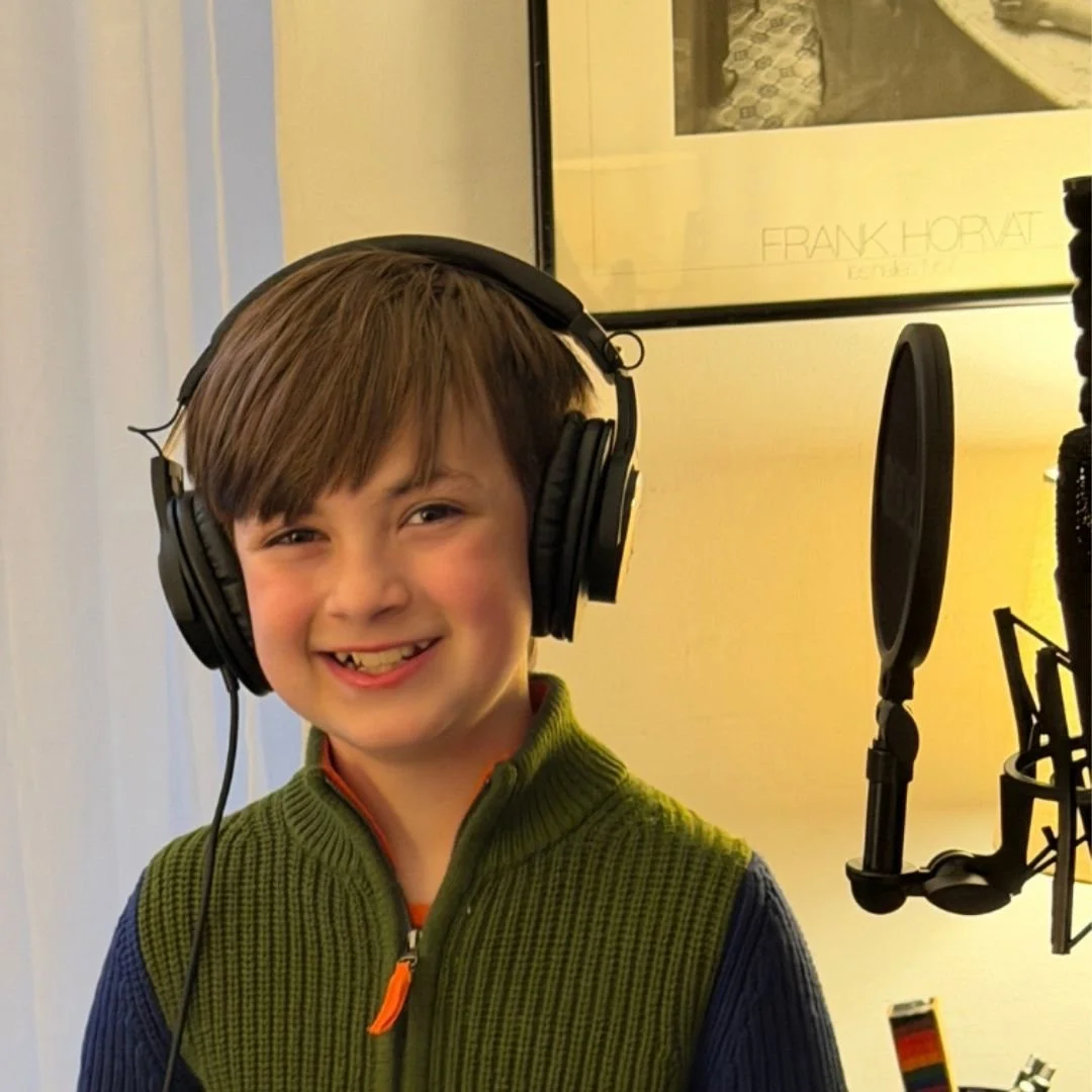 A close-up of a young boy wearing headphones and smiling while standing in front of a studio microphone and pop filter.