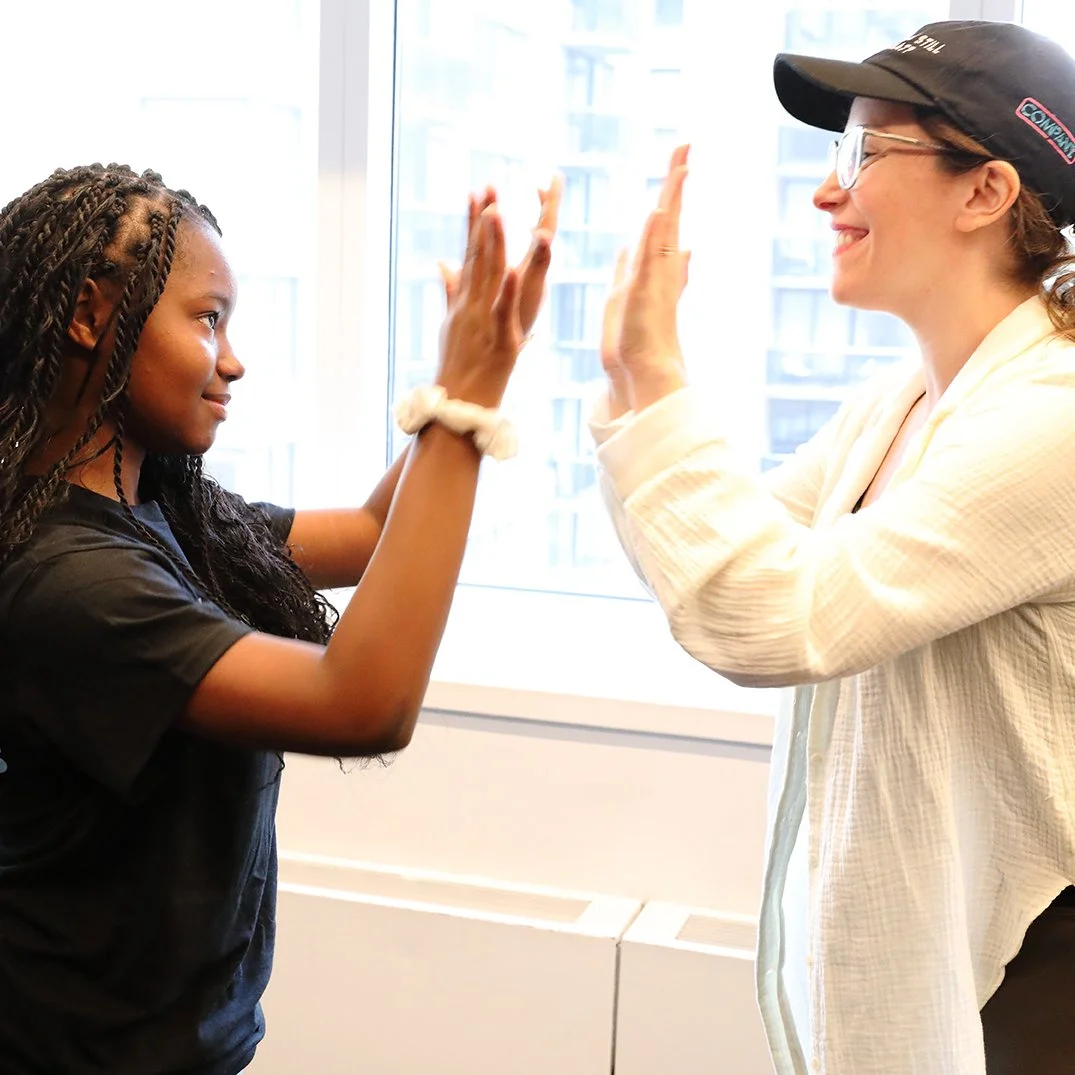 Detailed: A young girl with braided hair and a woman wearing glasses and a cap giving a double high-five in front of a bright window.