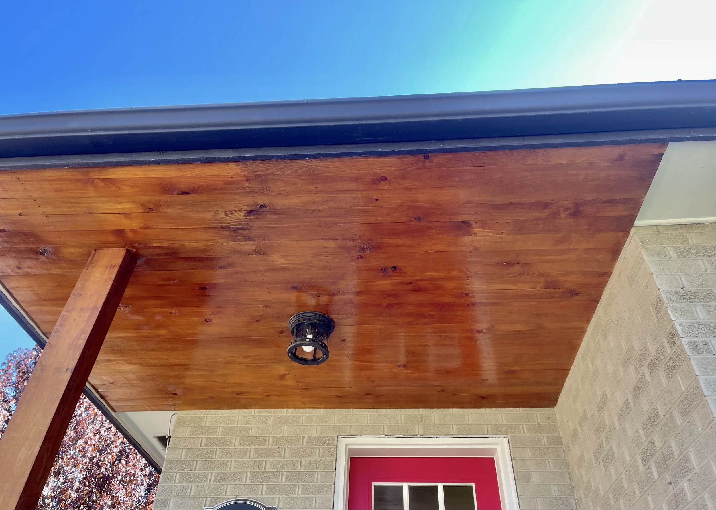 Exterior home improvement by Otto Custom Builders featuring a wood-paneled front porch ceiling, black light fixture, blue trim, brick walls, and a red entry door. Professional exterior remodeling, porch ceiling installation, woodwork, and painting.